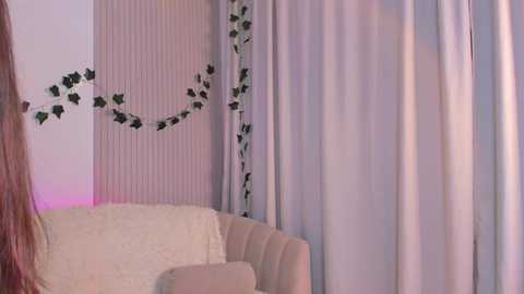 Media: Video of a cozy living room corner with a cream-colored sofa, adorned with a white fluffy blanket, and a green leaf garland on a vertical white panel. Soft pastel lighting adds a serene ambiance.