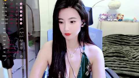 Media: A video of an Asian woman with long black hair and red lipstick, wearing a green floral dress, sitting in a chair with a teddy bear on a bed in the background.