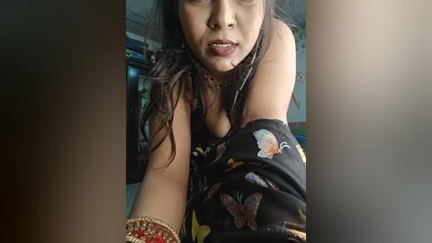 Media: Video of a South Asian woman with medium skin tone, wearing a black floral dress, gold bracelets, and dark lipstick, taken from a low angle. Background shows a cluttered room with green and blue objects.