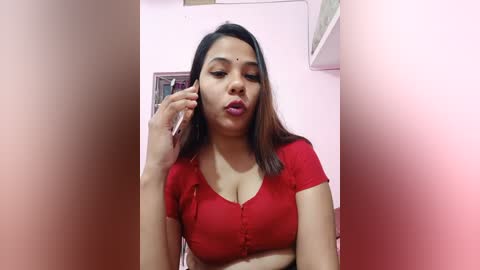 Media: Video of a South Asian woman with long dark hair and fair skin, wearing a red, short-sleeved, button-down top, making a pouty face while talking on a phone, in a pastel-colored room with shelves.