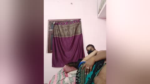 Media: Video of a woman in a traditional purple and green saree, lying on a bed in a simple, pastel-painted room with a purple curtain and a window.