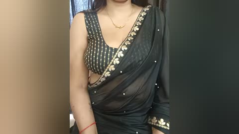 Media: Video of a woman wearing a black saree with gold floral embroidery, gold necklace, and red bracelet, standing indoors.