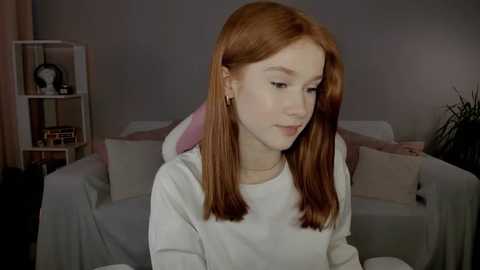 Media: A video of a fair-skinned young woman with long, straight auburn hair, wearing a white hoodie, sitting indoors in a cozy living room with a gray couch, bookshelves, and potted plants.