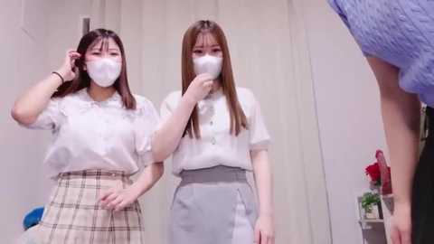 Media: Video of two Asian women in white masks, white blouses, and plaid skirts, standing indoors near a door. A man in blue clothing is partially visible.