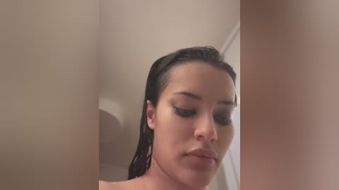 Media: Video of a young woman with medium skin tone, dark, slicked-back hair, and closed eyes, standing in a beige-tiled bathroom, wearing minimal makeup.