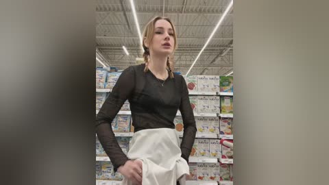 Media: Video of a slender, fair-skinned woman with blonde hair, wearing a sheer black top and white skirt, standing in a grocery store with brightly lit shelves full of colorful products.