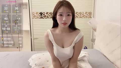 Media: Video of an East Asian woman with long brown hair, wearing a white off-shoulder top, sitting on a bed with cream sheets. Background shows a beige window with blinds, and a digital screen displaying time and weather.