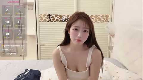 Media: Video of an East Asian woman with long brown hair and fair skin, wearing a beige tank top, sitting on a bed with white bedding. Background shows a closed white window blind and a calendar.