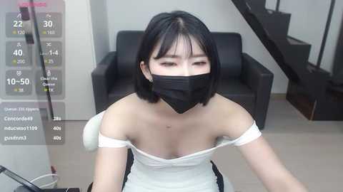 Media: Video of an East Asian woman with short black hair, wearing a white off-shoulder dress, black face mask, and a white headband, sitting on a chair in a modern living room with a black armchair and staircase.