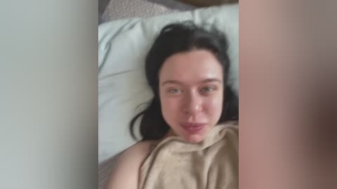 Media: A video of a young woman with fair skin and long black hair, lying on a bed with a beige blanket, wearing a light brown top.