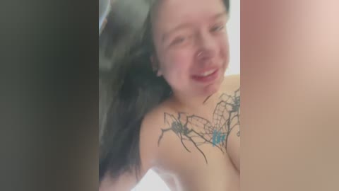 Media: Video of a fair-skinned woman with long black hair, smiling, showing a spider web tattoo on her shoulder, and a blue butterfly design on her chest.