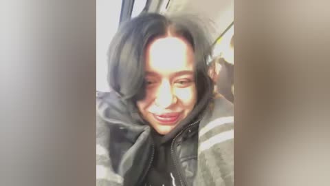 Media: Video of a smiling woman with dark hair, wearing a gray puffer jacket, seated in a car with sunlight streaming in.