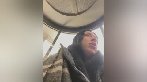 Media: Video of a young man with fair skin, wearing a black hooded jacket and headband, looking up with a surprised expression, standing inside a bus.