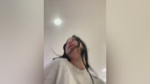 Media: A blurred video of a young woman with long, dark hair, wearing a white t-shirt, standing against a plain white wall with a ceiling light.