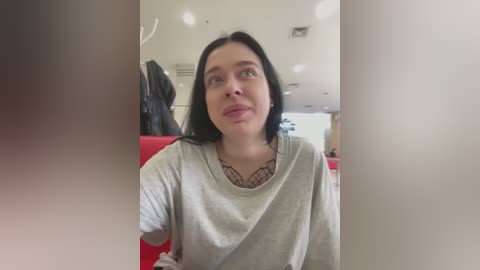 Media: Video of a young woman with fair skin, black hair, and a light grey t-shirt, sitting on a red couch in a modern, well-lit room with white walls and a hanging jacket.