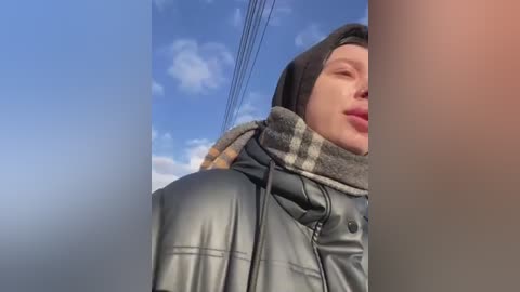 Media: Video of a fair-skinned woman with long dark hair, wearing a black leather jacket, gray scarf, and a beige plaid scarf, standing against a blue sky with scattered clouds and power lines in the background.