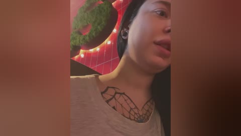 Media: Video of a young woman with medium skin tone, wearing a beige shirt, black necklace, and headphones, set against a dimly lit background with a red and green patterned wall and a circular light fixture.