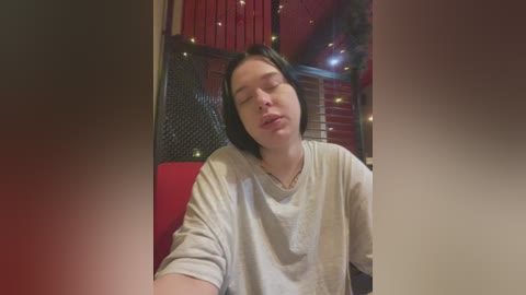 Media: A video of a fair-skinned, young woman with short black hair, wearing a light gray t-shirt, resting her head on her hand in a dimly lit room with red curtains and string lights.