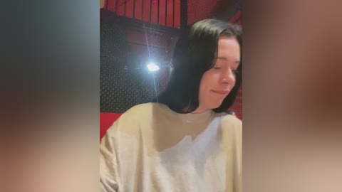 Media: Video of a young woman with shoulder-length black hair, wearing a light-colored sweater, smiling, with a bright light illuminating her face. Background features red and black walls with a mesh texture.