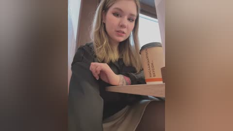 Media: Video of a young Caucasian woman with light skin and shoulder-length blonde hair, wearing a black sweater, leaning against a wooden counter in a cafe, holding a Starbucks cup.