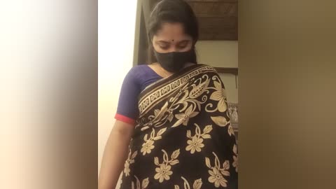 Media: Video of a woman with medium skin tone, wearing a black sari with white floral patterns and red trim, a black mask, and a forehead bindi, standing indoors, with blurred background.