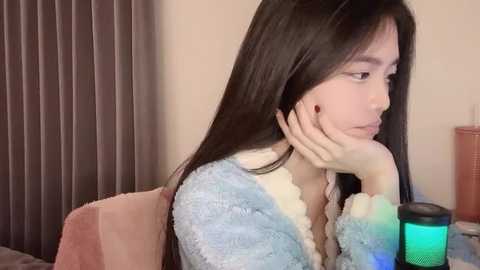 Media: Video of a young Asian woman with long, straight black hair, wearing a light blue, fluffy robe, sitting pensively with her chin on her hand, next to a lit teal lamp on a bed with beige curtains in the background.