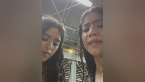 Media: Video of two young women with long dark hair and light skin, looking distressed, standing in a dimly lit, industrial-looking room with metal walls and a high ceiling.