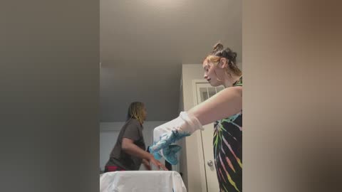 Media: Video of two women in a sparsely furnished room, one in a tie-dye shirt and the other in a black top, applying blue painter's tape to a white table.