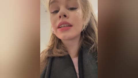 Media: Video of a young Caucasian woman with fair skin, light brown hair, and closed eyes, wearing a gray cardigan over a pink shirt, taken indoors with a soft-focus background.