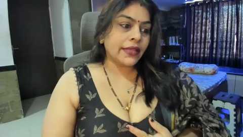 Media: Video of a South Asian woman with medium brown skin, wearing a black top with butterfly pattern, sitting in a modern living room with wooden floors, dark curtains, and a beige sofa.