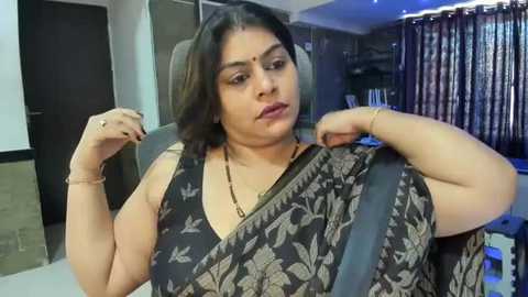 Media: A video of a middle-aged Indian woman with medium skin tone and shoulder-length dark hair, wearing a black floral sari, adjusting a seatbelt in a modern, dimly lit living room with dark curtains and minimalist decor.