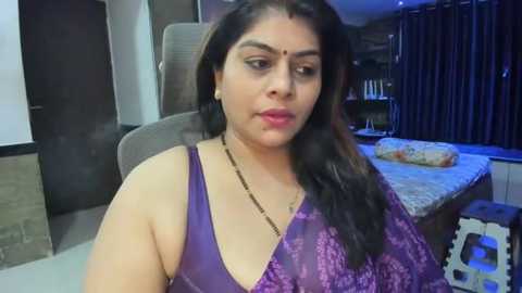 Media: Video of a South Asian woman with medium brown skin, wearing a purple, semi-sheer lace dress, seated indoors, surrounded by modern decor with dark curtains and a white couch.