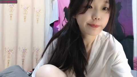 Media: Video of a young Asian woman with long black hair, light skin, and a slight smile, wearing a white robe, holding a baby. Background features a pink and purple wall and white curtains with floral patterns.