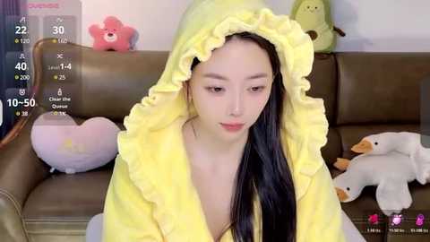 Media: Video of a young Asian woman with long black hair, wearing a yellow hoodie with frills, sitting on a brown leather couch. Background features plush toys and a digital screen showing a camera view.