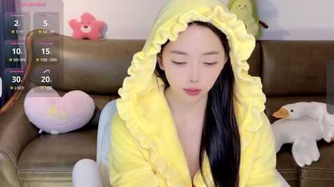 Media: Video of a young East Asian woman with long black hair, wearing a yellow hooded bathrobe, sitting on a green couch with plush toys, including a pink star, a yellow duck, and a white rabbit.