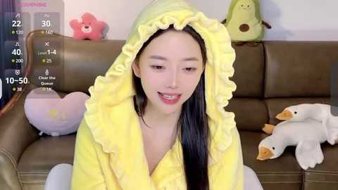 Media: Video of an East Asian woman in a yellow, frilled bathrobe, smiling on a brown couch with plush toys.