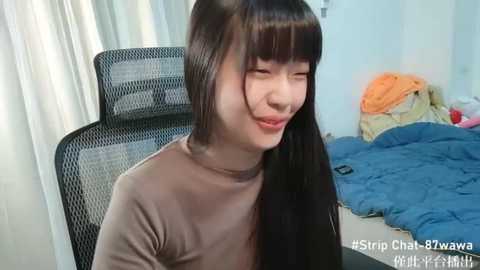 Media: A video of a young Asian woman with long, straight black hair and fair skin, wearing a beige turtleneck sweater, sitting on a black mesh chair in a messy bedroom.