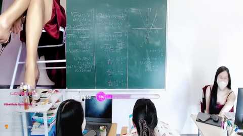 Media: Video of an Asian woman in a red dress, sitting on a chair, writing on a green chalkboard with complex equations. Two other women with long hair, one in a black dress, the other in a white top, sit at a desk studying.