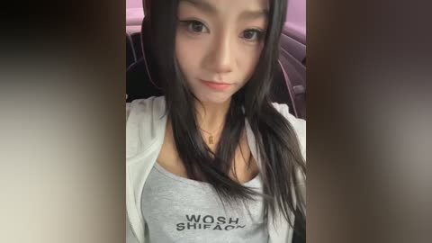 Media: Video of a young Asian woman with long black hair, fair skin, and a petite build. She wears a grey tank top with the words \"WOSH SHIPO\" in black. She sits in a car with a blurred background.