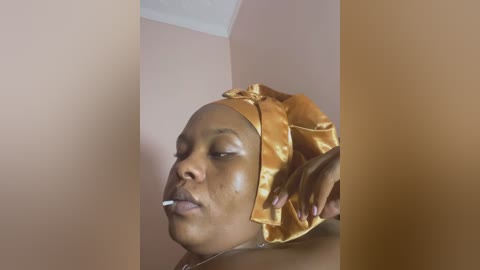 Media: Video of a dark-skinned woman with a gold headwrap, cigarette in mouth, and a hand adjusting her wrap, in a pale pink room with a white ceiling.