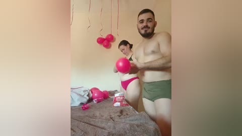 Media: Video of a smiling, bearded man in green boxer briefs and a woman in red lingerie, holding pink boxing gloves in a bedroom with balloons, bed, and snacks.