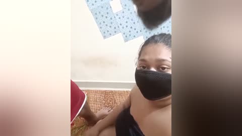 Media: A video shows an Asian woman with a black face mask and brown hair tied back, sitting on a wicker chair with a red shirt, in a room with blue patterned paper on the wall.