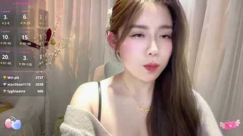 Media: Video of an East Asian woman with long brown hair, fair skin, and red lipstick, wearing a beige off-shoulder sweater, standing against a white curtain background.