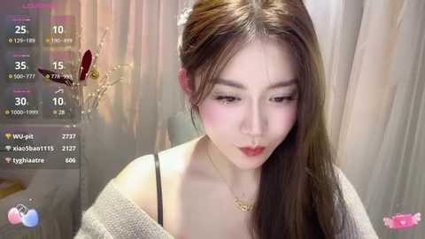 Media: A video of an East Asian woman with long brown hair, wearing a gray off-shoulder sweater, and a gold necklace, set against a blurred, beige background.