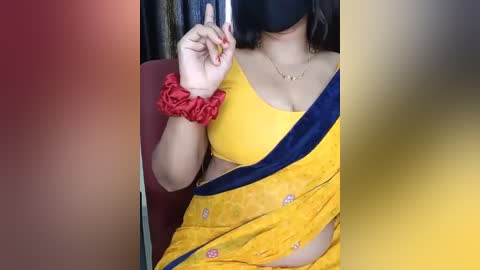 Media: Video of a South Asian woman in a yellow saree with a blue border, wearing a red scrunchie and black face mask, holding a phone. Background is blurred.