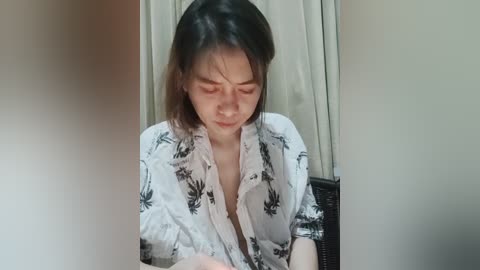 Media: Video of a young Asian woman with shoulder-length dark hair, wearing a white shirt with black floral patterns, sitting indoors. She has a contemplative expression, with a soft, neutral background.