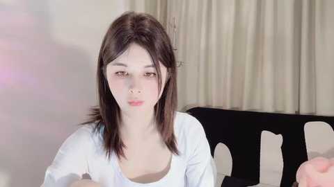 Media: Video of a young Asian woman with straight, shoulder-length dark brown hair, wearing a white top, seated indoors with beige curtains in the background.