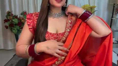 Media: Video of a South Asian woman in a vibrant red saree with intricate gold embroidery, wearing matching jewelry, including a choker, bangles, and earrings. She has long brown hair and a confident smile.