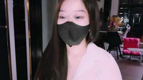 Media: Video of a young Asian woman with long black hair, wearing a black face mask and white shirt, in a modern indoor setting with pink chairs and floral arrangements.