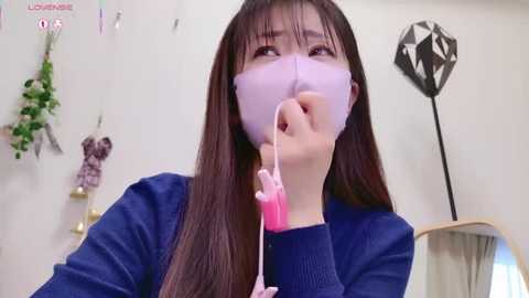 Media: Video of an Asian woman with long brown hair wearing a lavender face mask, blue sweater, and pink hand sanitizer, in a brightly lit room with minimalistic decor.